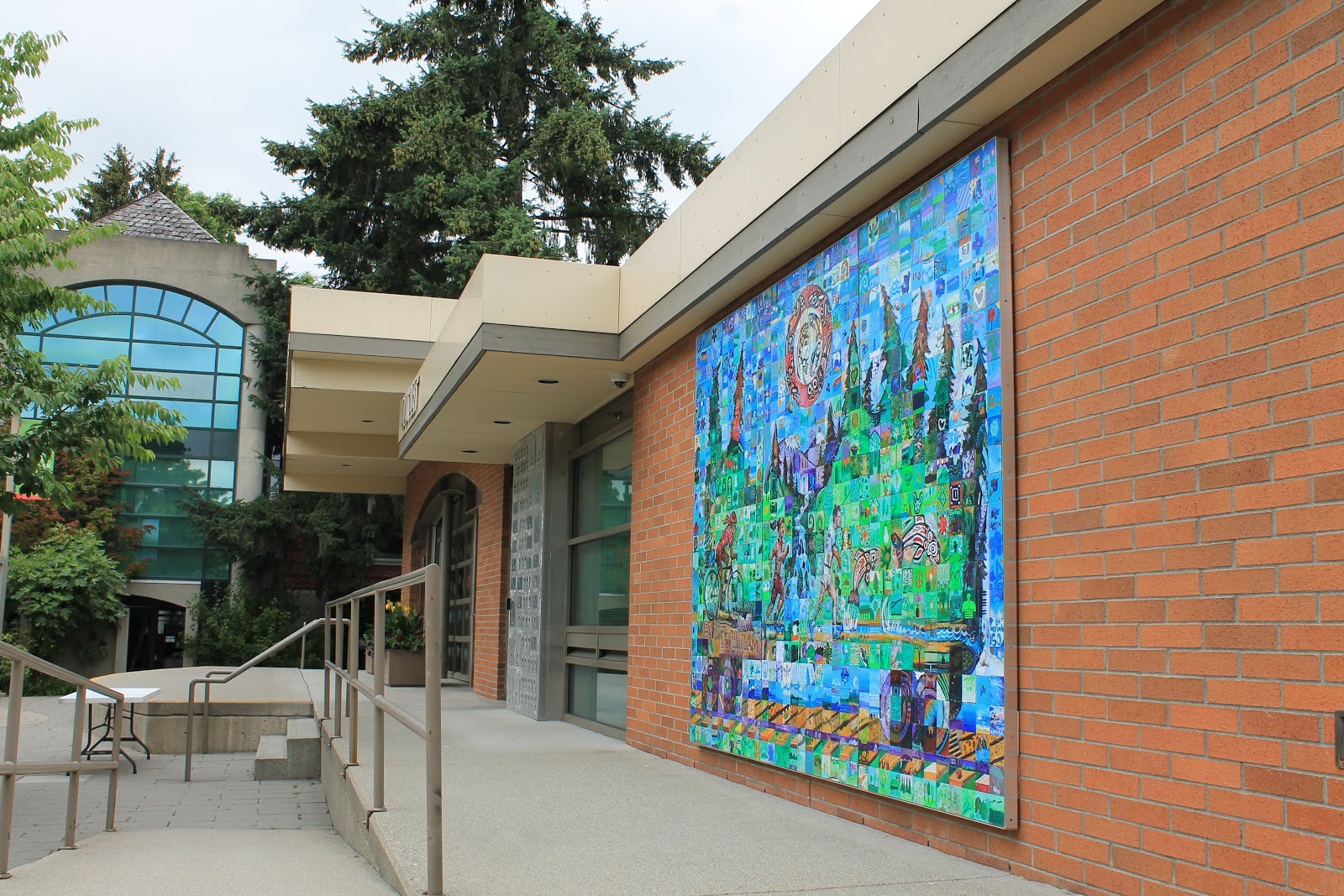 A photo of the back of the Leigh Square Community Arts Village building. There is a wheelchair ramp, a door, and a colourful mosaic mural about Port Coquitlam.