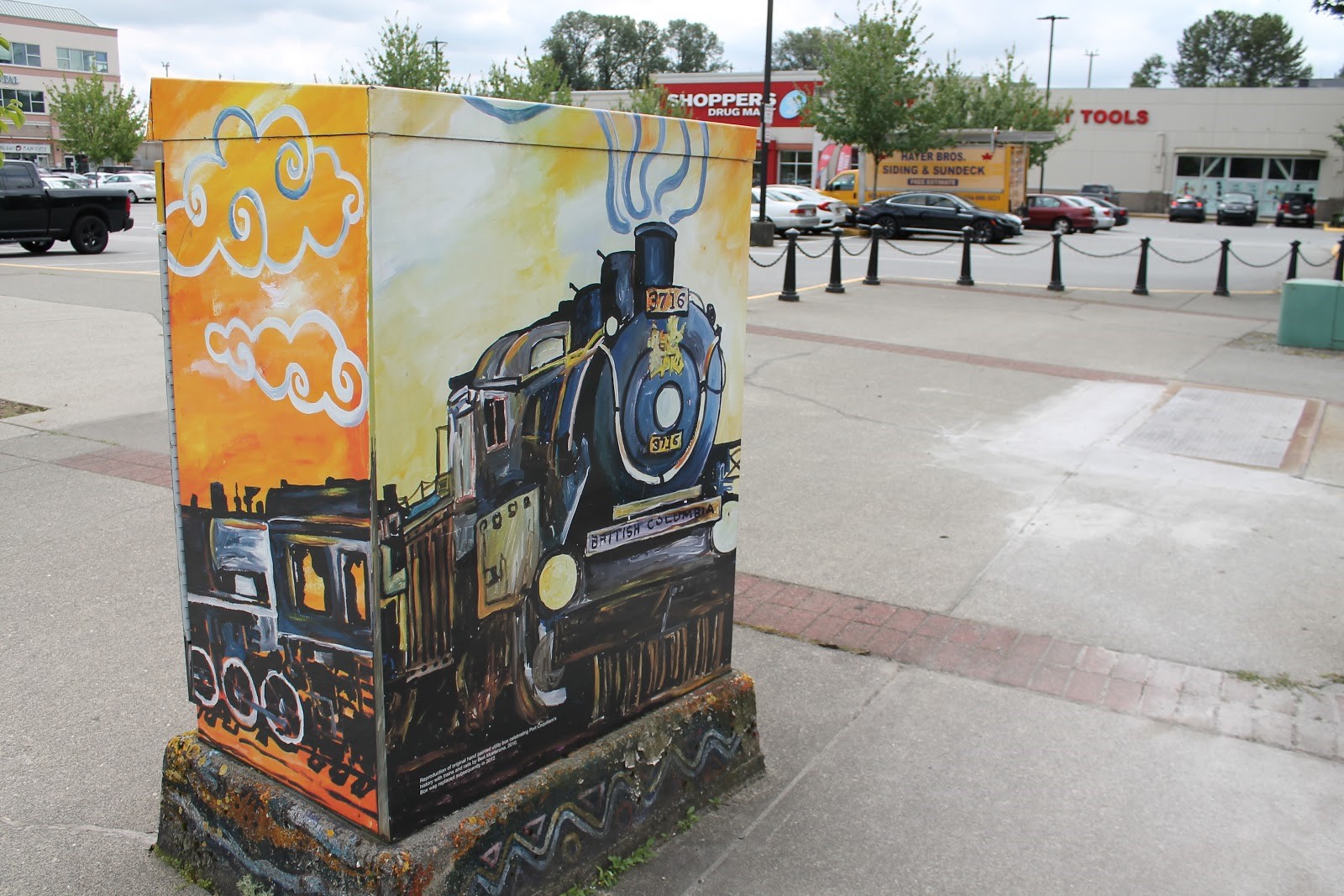 A photograph of an electrical box on the sidewalk near a shopping centre parking lot with a painting of a train on it.