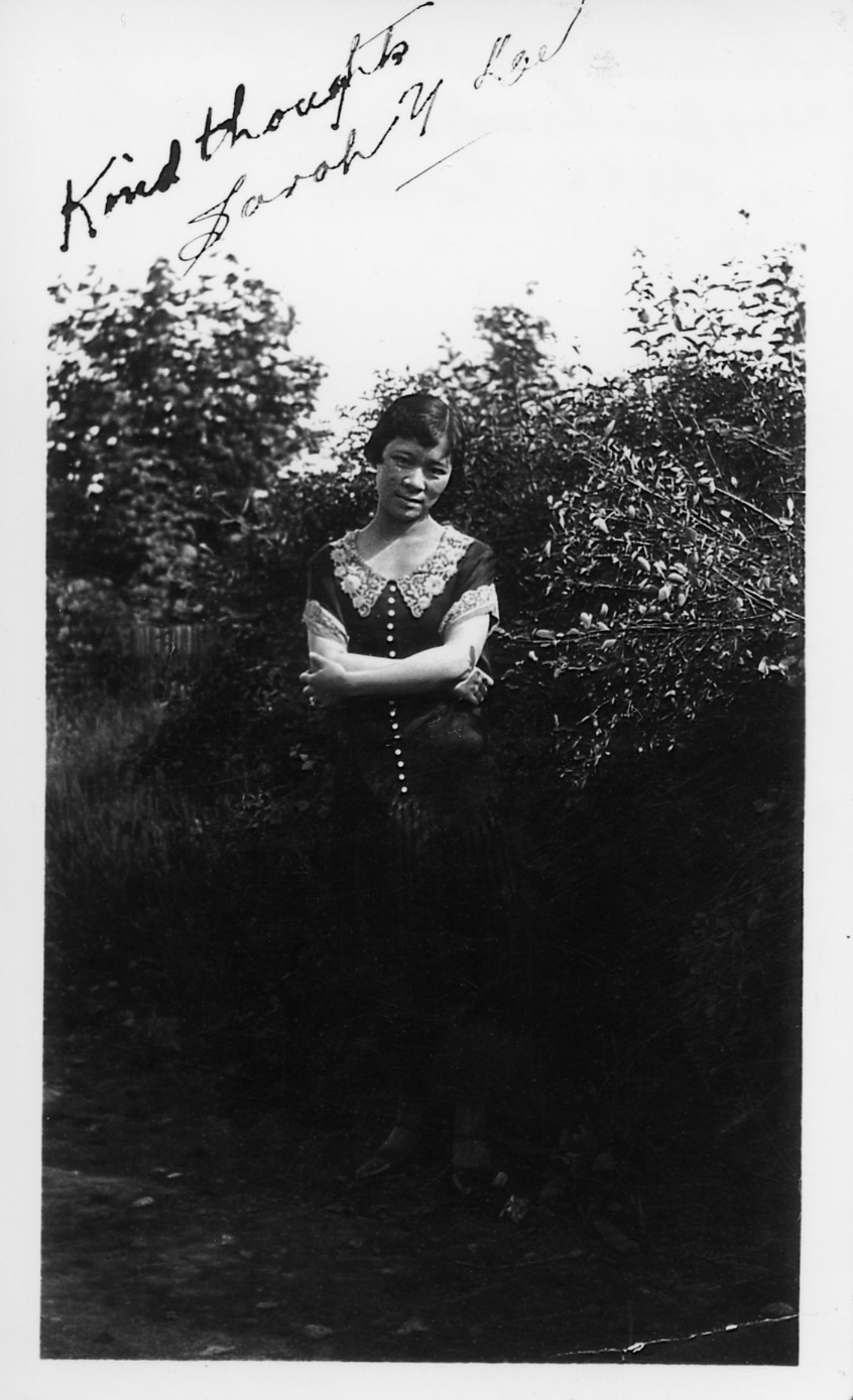 A black and white photo of a young girl wearing a black dress with white collar posing in front of a tree