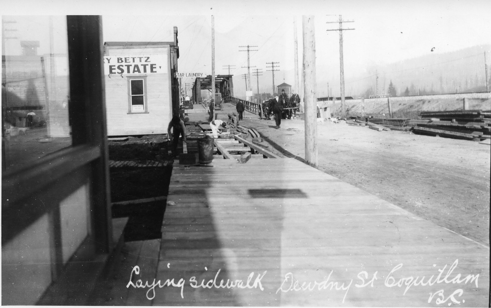 A black and white photo of a wooden sidewalk and dirt road with store fronts on the left and bridge in the distance