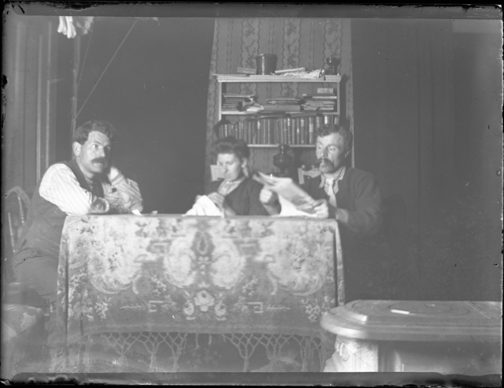 Candid black and white photo of two men and a woman reading and sewing at a table in a dark house.