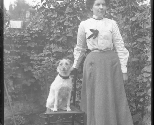 Black and white photo of a woman in period clothing standing in a garden. A small dog sits on a chair next to her
