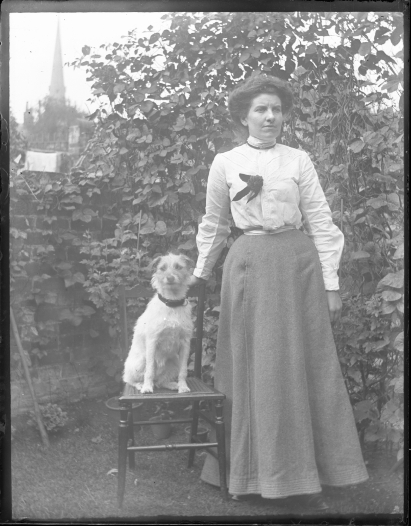 Black and white photo of a woman in period clothing standing in a garden. A small dog sits on a chair next to her