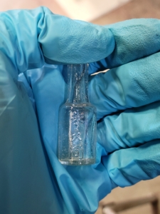 A gloved hand holds a very small, clear glass bottle embossed with Chinese characters