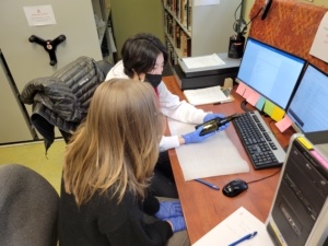 Two gloved people sit at a desk, one holds a glass bottle that they are looking at