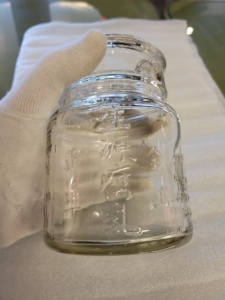 A gloved hand holds a short, clear glass bottle with embossed Chinese characters