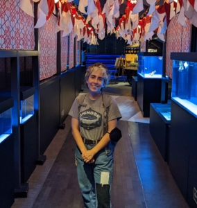 Paisley standing in a hall full of lanterns at an aquarium in Japan. She is wearing a grey shirt with a Chinook salmon on it, and jeans.