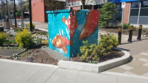 A photo of an electrical box with a stylized art piece of red salmon in swirling turquoise water on the edge of a garden. 